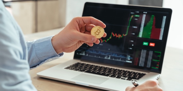 Eine Person hält eine Bitcoin-Münze vor einem Laptop mit Kurscharts und Handelsdaten. Neutrales Krypto-Motiv im Kontext Bitcoin kaufen: Wie seriös ist Bitpanda?